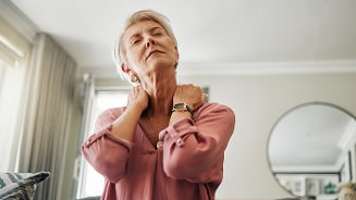 Persoon in roze blouse met handen op de nek, in een woonkamer.