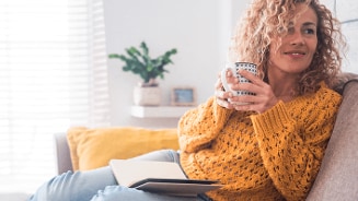 Vrouw in een gele trui met een kopje koffie op de bank, naast haar ligt een geopend notitieboek.
