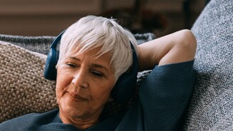 Ouder persoon met grijs haar ontspant op een bank en draagt koptelefoon.