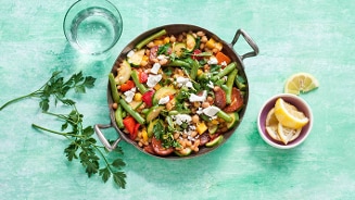 Gekleurde groenteschotel met bonen en feta op een groene achtergrond, geserveerd met een glas water en partjes citroen.