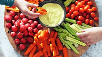 Groenteplank met hummusdip, omringd door radijsjes, wortels, bleekselderij, snoeptomaten en zoete puntpaprika's.