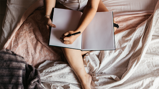 Persoon schrijft in een notitieboek terwijl hij op bed zit.