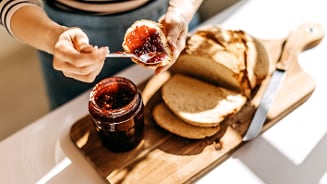 Persoon smeert jam op een sneetje brood naast een snijplank met gesneden brood en een pot jam.
