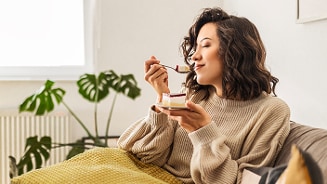 Vrouw in een beige trui, zittend op een bank en genietend van een dessert met een groene plant op de achtergrond.