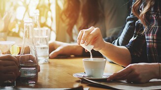 Persoon die suiker toevoegt aan een kop koffie in een café-omgeving.