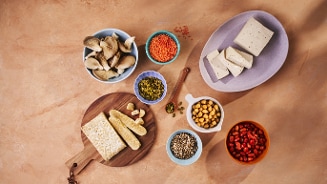 Verschillende plantaardige ingrediënten zoals tempeh, tofu, kikkererwten, linzen, zonnebloempitten, granaatappel en oesterzwammen op een houten snijplank en in kleurrijke kommen.