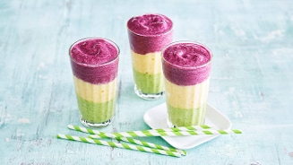 Drie gelaagde smoothies in glazen met paarse, gele en groene lagen, op een lichtblauwe achtergrond met papieren rietjes.