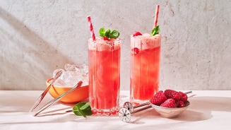 Twee glazen frambozenlimonade met ijs en muntblaadjes, naast een schaaltje verse frambozen.