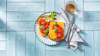 Twee volkoren boterhammen met kaas, tomaat en verse basilicum op een bord, naast een glas thee.