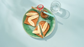 Twee sneetjes toast met plakjes banaan en een potje pindakaas op een groen bord, met een glas water erbij.