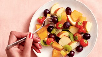 Bord met vers fruitsalade bestaande uit aardbeien, druiven, appelpartjes, kiwi en meloen, met een hand die een vork vasthoudt.