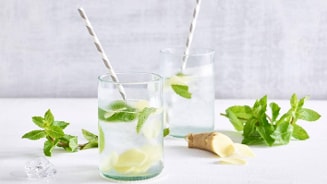 Twee glazen met verfrissend water, schijfjes gember, munttakjes en papieren rietjes.