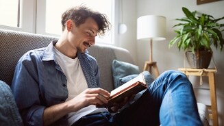 Jongere leest een boek op de bank in een gezellige woonkamer met kamerplant en vloerlamp