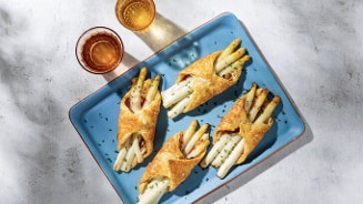 Bladerdeegpakketjes met witte asperges en chorizo op een blauw bord, met twee glazen amberkleurige drank.