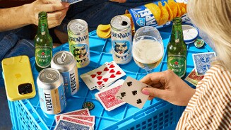 Spelers zitten rond een blauwe krat en spelen een kaartspel met verschillende open kaarten, omringd door blikjes Vedett bier, Brouwenwolf witbier, flessen Heineken 0.0 witbier, een glas bier, chips en een telefoon.