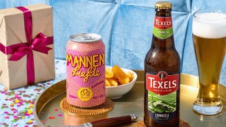 Blik Mannenliefde bier en fles Texels Springtij speciaalbier op een rond dienblad met een glas bier, een bakje chips en een cadeau met roze strik op de achtergrond.