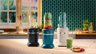 Drie blue blenders op een aanrecht, met in het midden een blender gevuld met vers fruit en groenten. Rechts staat een glas met groene smoothie en een bordje met vers fruit. De keuken op de achtergrond heeft een groene tegelmuur en een raam.