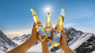 Drie handen houden flessen Corona Cero vast met een schijfje limoen in de flessenhals, met een besneeuwd berglandschap en een heldere blauwe lucht op de achtergrond.