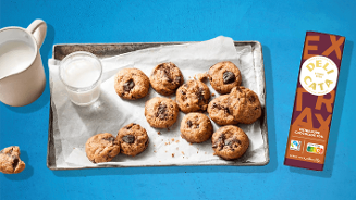 Chocoladekoekjes met stukjes pure chocolade liggen op een bakplaat met een glas melk erbij, naast een reep Delicata extra pure chocolade 70%.