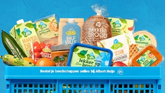 Blauwe Albert Heijn boodschappenkrat gevuld met diverse Albert Heijn huismerkproducten, waaronder groenten, brood, zuivel, hummus, crackers en vleeswaren, tegen een blauwe achtergrond. Tekst op de krat: "Bestel je boodschappen online bij Albert Heijn".