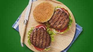 Twee gegrilde hamburgers met sla, tomaat en ui op een houten plank, naast een sesambroodje.