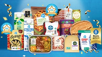 Een assortiment van Albert Heijn producten, waaronder zuivel, groente, kant-en-klaar maaltijden, kaas, brood en diepvriesproducten, gepresenteerd tegen een blauwe achtergrond met feestelijke confetti. Meerdere producten hebben een keurmerk of prijs gewonnen, zoals de "VriesVers Winnaar 2025" en "Beste getest" labels.