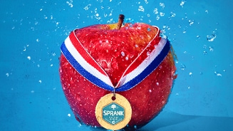 Rode appel met een Nederlandse medaille en het label 'Sprank', omgeven door waterdruppels op een blauwe achtergrond.