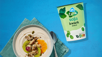 Schaal met ongezoete AH Terra soja kwark, gegarneerd met kiwi, mandarijn, druif en noten, naast een verpakking van AH Terra soja kwark op een blauwe achtergrond.