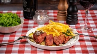 Bord met gegrilde vleesstukken en frietjes op een rood-wit geruite tafelkleed, omgeven door een glas rode wijn en een kom groene sla.