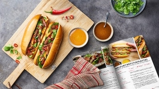 Banh mi met biefstukpuntjes op een houten snijplank, omringd door sauzen en een open kookboek.