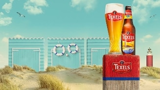 Texels Skuumkoppe 0.0 speciaalbier flesje en glas op een strand met strandhuisjes en vuurtoren op de achtergrond
