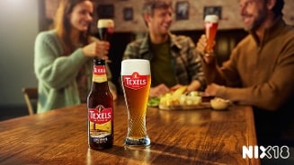 Fles en glas Texels Skuumkoppe bier op een houten tafel, met op de achtergrond drie vrienden die proosten in een café. NIX18 logo rechtsonder in beeld.