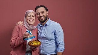 Twee mensen met onscherpe gezichten, gekleed in een roze blazer en blauw overhemd, houden een bord met eten en een boek vast tegen een rode achtergrond.