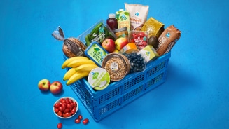 Blauwe boodschappenkrat gevuld met diverse AH-producten, waaronder bananen, appels, cherrytomaten, blueberry, broden, muesli en havermoutdrink.