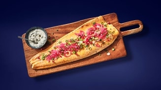 Gegratineerde visfilet op een houten serveerplank, gegarneerd met rode ui en groene kruiden, met een schaaltje saus ernaast.