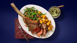 Tomahawk steak geserveerd met gebakken aardappeltjes, sperziebonen met spek en kruidenboter op een witte schaal, tegen een blauwe achtergrond.