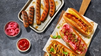 Gegrilde worsten en hotdogs met verschillende toppings, geserveerd met bakjes ingelegde uien en ketchup op een donkere ondergrond.