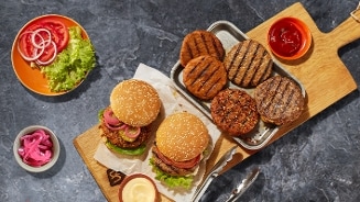 Twee vegetarische hamburgers op een broodje met sla, tomaat en ingelegde ui, geserveerd op een houten plank met diverse gegrilde vegaburgers, een bakje ketchup, een schaaltje ingrediënten en een kommetje mayonaise.