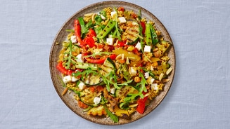 Bord met mediterrane couscoussalade, inclusief groenten zoals paprika, courgette, en rucola, verrijkt met stukjes feta en amandelen.