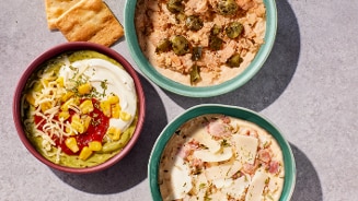 Drie kleurrijke kommen met verschillende dips en gerechten, waaronder een Mexicaanse dip met mais, guacamole en zure room, een witte dip met kaas en ham, en een kom met een tonijn- en groentemix, naast enkele stukjes flatbread op een stenen ondergrond.