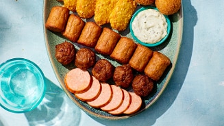 Schaal met verschillende borrelhapjes zoals gesneden worst, gehaktballetjes, mini loempia's, kaasbroodjes, saus in een bakje en een glas water op een blauwe ondergrond.
