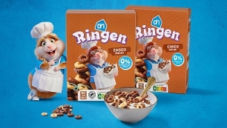 Twee pakken AH Ringen Choco smaak ontbijtgranen met een cartoonhamster in bakkerstenue, een kom ontbijtgranen met melk en losse granen op een blauwe achtergrond.