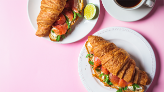 Croissant belegd met zalm, roomkaas en rucola op een bord met een halve limoen en een kop koffie op een roze ondergrond