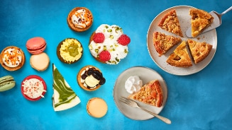 Diverse selectie van gebak, macarons en taartpunten op een blauwe ondergrond, met onder andere een appelkruimeltaart, fruitgebakjes, cheesecake en slagroom.