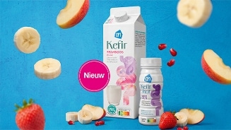 Twee AH Kefir producten (framboos en multivruchten shot) omringd door plakjes banaan, stukjes appel, aardbei en granaatappelpitten op een blauwe achtergrond, met een roze label "Nieuw".