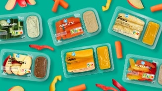 Assortiment van Albert Heijn snackgroenten en fruit in plastic verpakkingen, waaronder komkommer, worteltjes, ananas, rode appeltjes, mango en paprikamix met bijpassende dips, op een turquoise achtergrond.