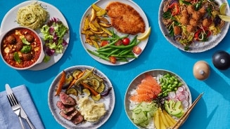 Vijf borden met verschillende maaltijden op een blauwe ondergrond, waaronder pasta met gehaktballen, schnitzel met groenten, een Aziatische maaltijd met gefrituurde stukjes, een gerecht met gebakken vlees en aardappelpuree, en een pokébowl met zalm, avocado, radijs en edamame.