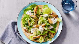 Wrap met blaadjes sla, stukjes gerookte makreel, komkommer en cottage cheese, geserveerd op een blauw bord met een glas water erbij.
