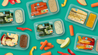 Assortiment van Albert Heijn snackgroenten en fruit in plastic verpakkingen, waaronder komkommer, worteltjes, ananas, rode appeltjes en paprikamix met bijpassende dips, op een turquoise achtergrond.