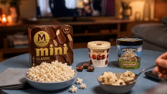 Een gezellige setting met een schaaltje popcorn, Magnum Mini Classic ijs, Franui melkchocolade met frambozen, en Ben & Jerry’s Cookie Dough ijs op een tafel, met iemand die een hapje ijs neemt.